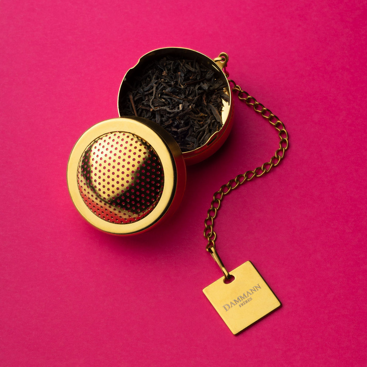 Dammann Frères gold-finish tea ball with loose tea on a pink background