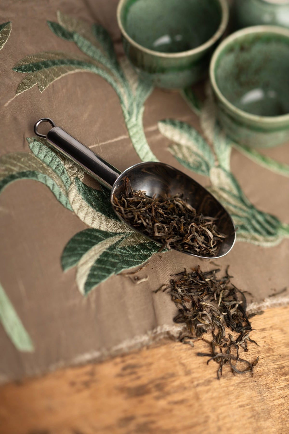 Cuillère doseuse à thé en acier inoxydable remplie de feuilles de thé blanc en vrac, sur un tissu à motif botanique
