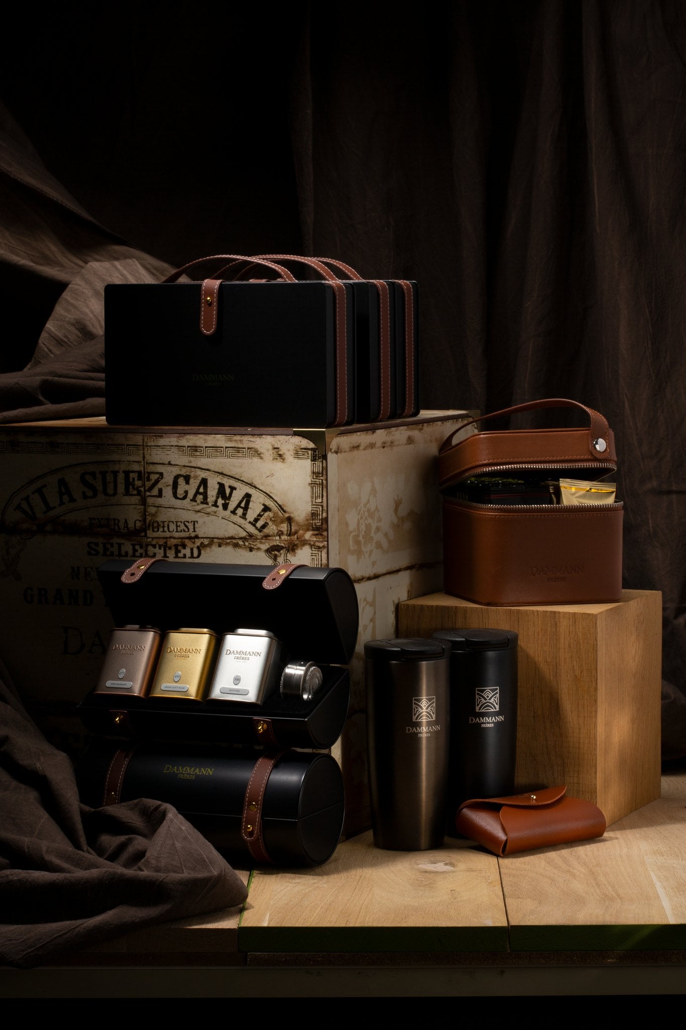 Dammann Frères Merveilleux black strapped case open with three tea tins and an infuser, beside a vintage tea chest