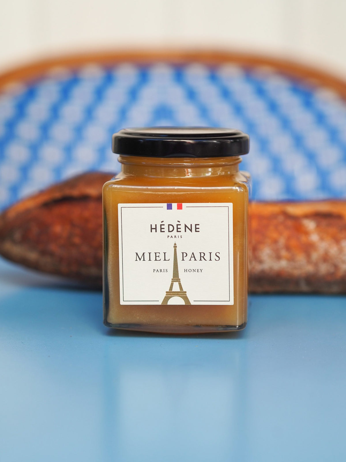 Hédène Paris' Honey jar with its golden Eiffel Tower label in front of a baguette and a bistro chair