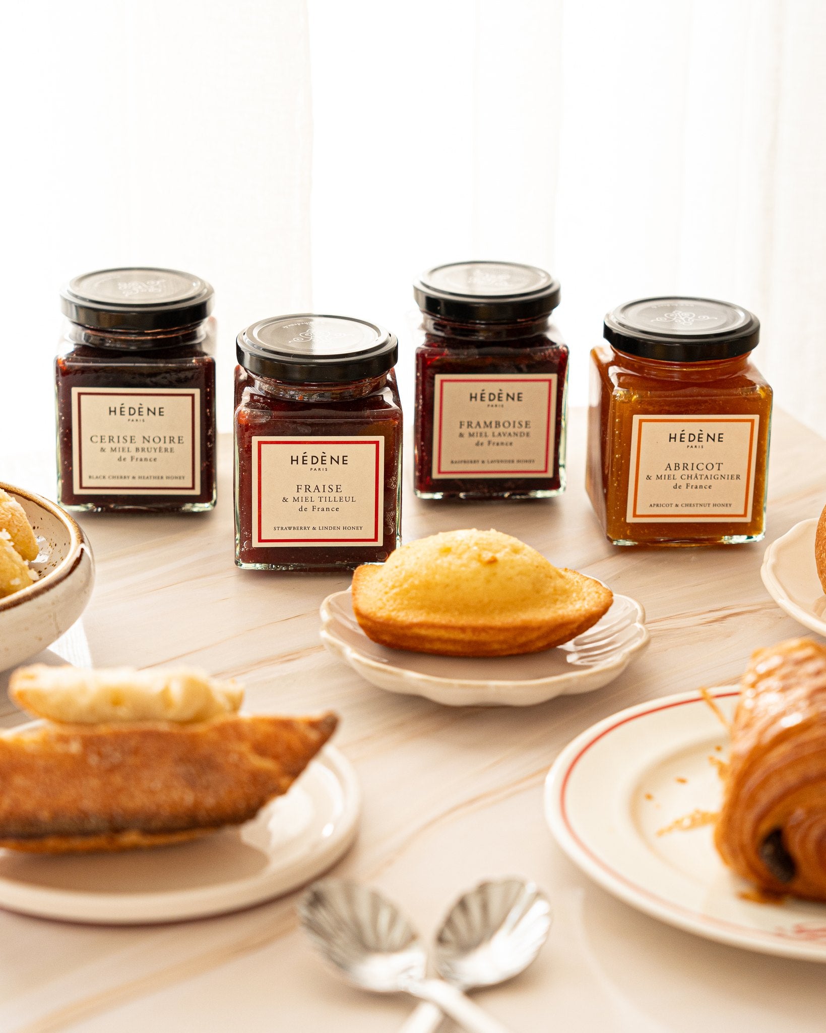 Les quatre confitures au miel Hédène côte à côte, avec des madeleines